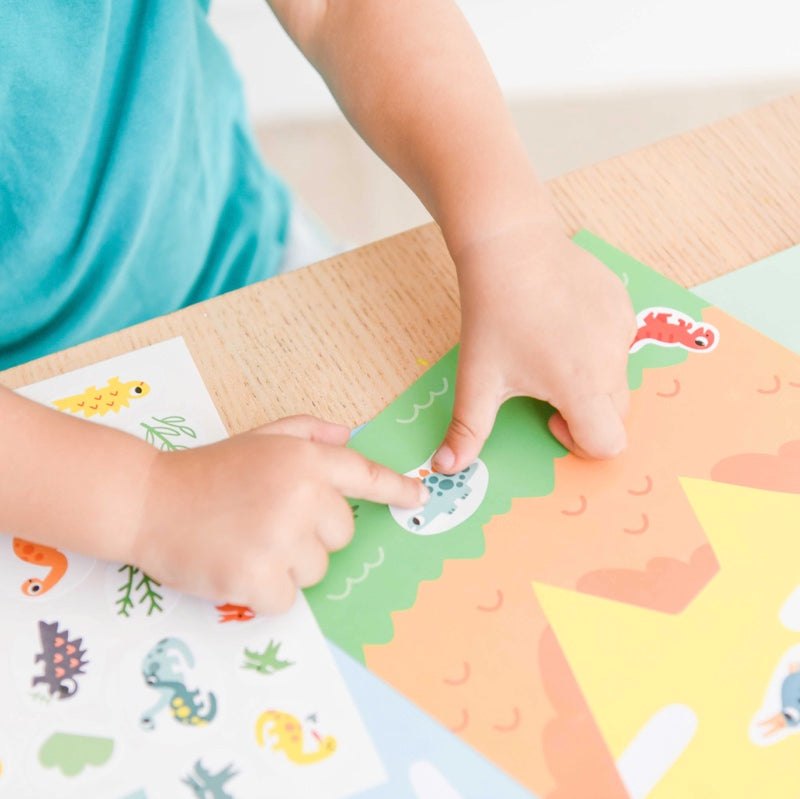 Child playing with Poppik Moje prve nalepnice, using colorful movable stickers on a large illustrated sheet.