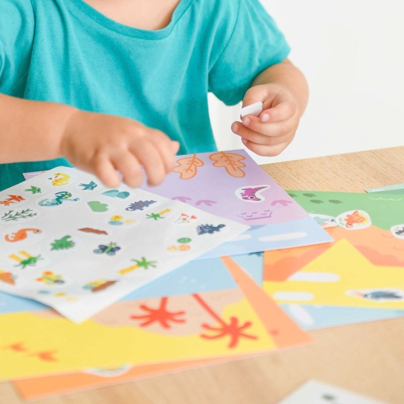 Child using Poppik Moje prve nalepnice with colorful sticker sheets on a table.