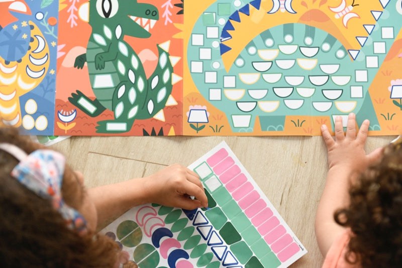 Colorful dinosaur sticker panorama with children assembling pieces on a large sheet, enhancing creativity and fine motor skills.